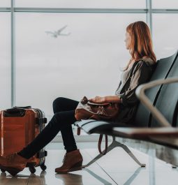 Tranquil woman is sitting on bench with luggage in lounge zone. She is looking through window at flying plane while waiting for her flight