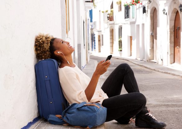 A woman sits on the sidewalk with her bags listening to music on her phone in a small Mediterranean-looking street