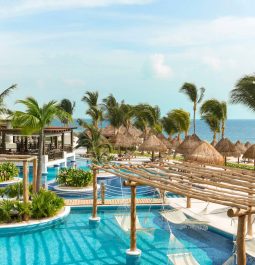 A luxurious tropical resort with a serene blue pool, surrounded by lush palm trees, thatched-roof cabanas, and hammocks. In the background, there's a calming view of the ocean under a clear blue sky.